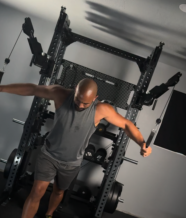 A man in a gray tank top and shorts exercising on a black multi-functional home gym cable machine with a power rack behind him.
